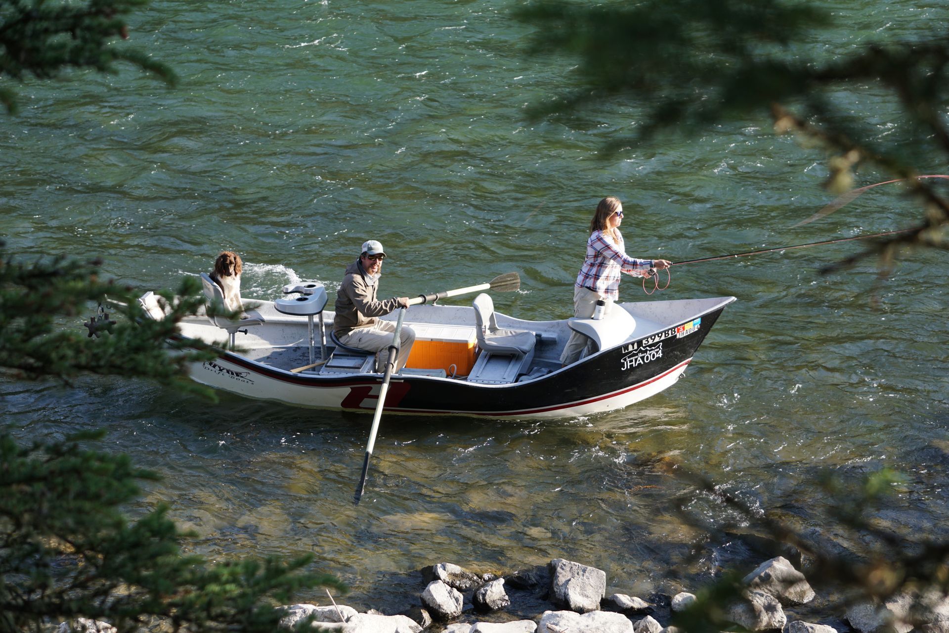 Three people in a small rowboat on a clear green river, one standing and rowing near rocky shore