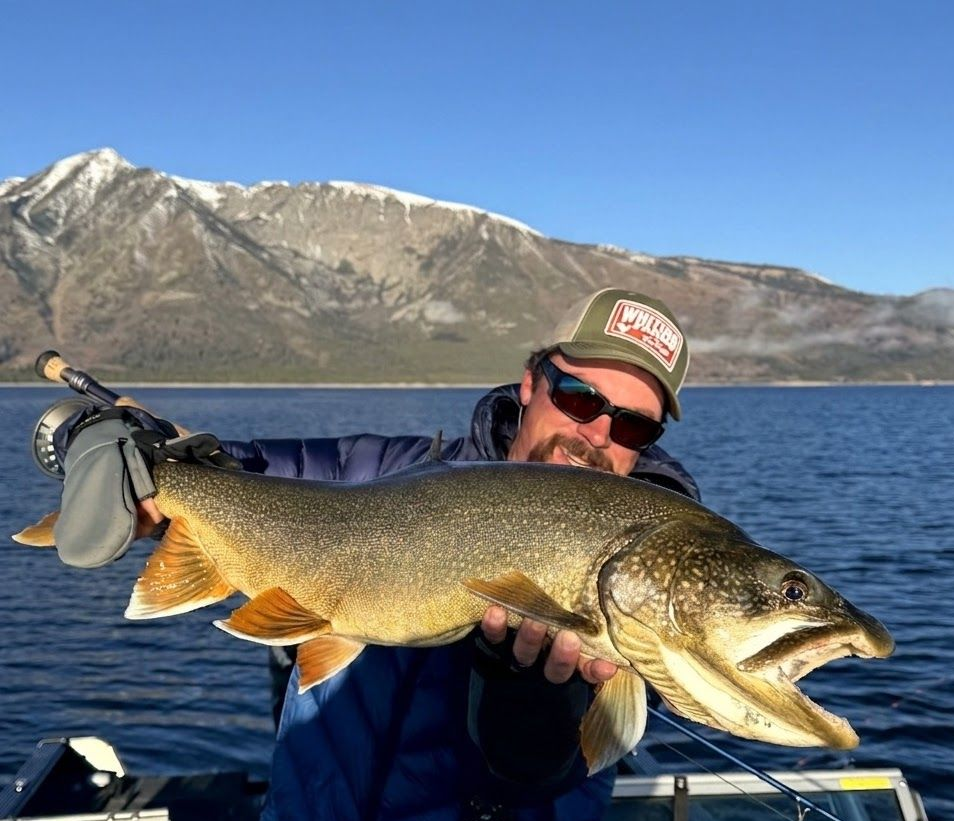 Angler holding a large yellow-spotted fish by a river, smiling in camo gear and sunglasses.