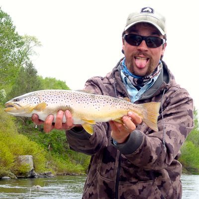Angler holding a large yellow-spotted fish by a river, smiling in camo gear and sunglasses.