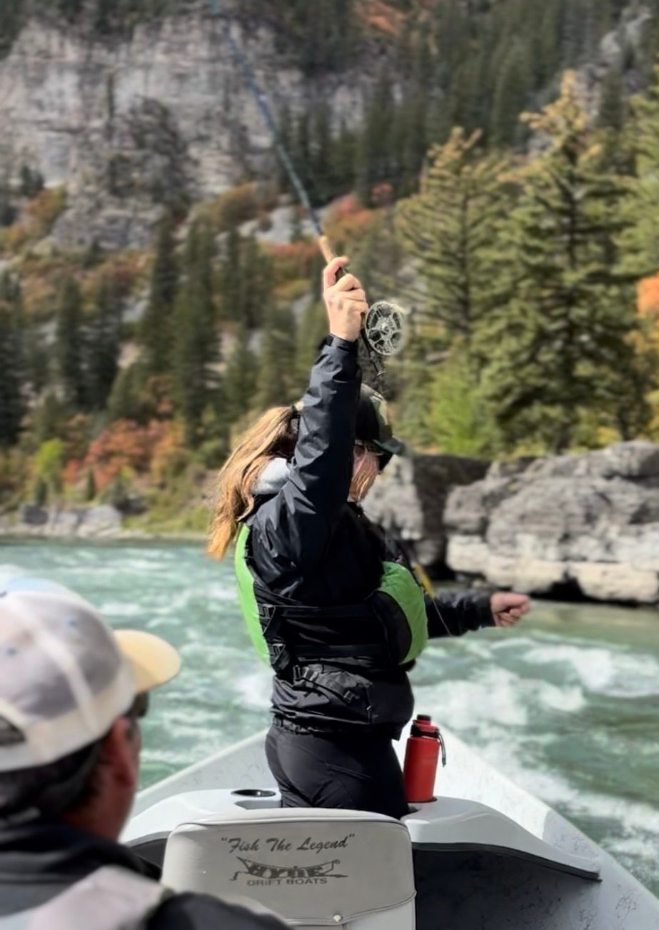 Person in a boat on a turquoise river, pointing upward near rocky cliffs and pine trees