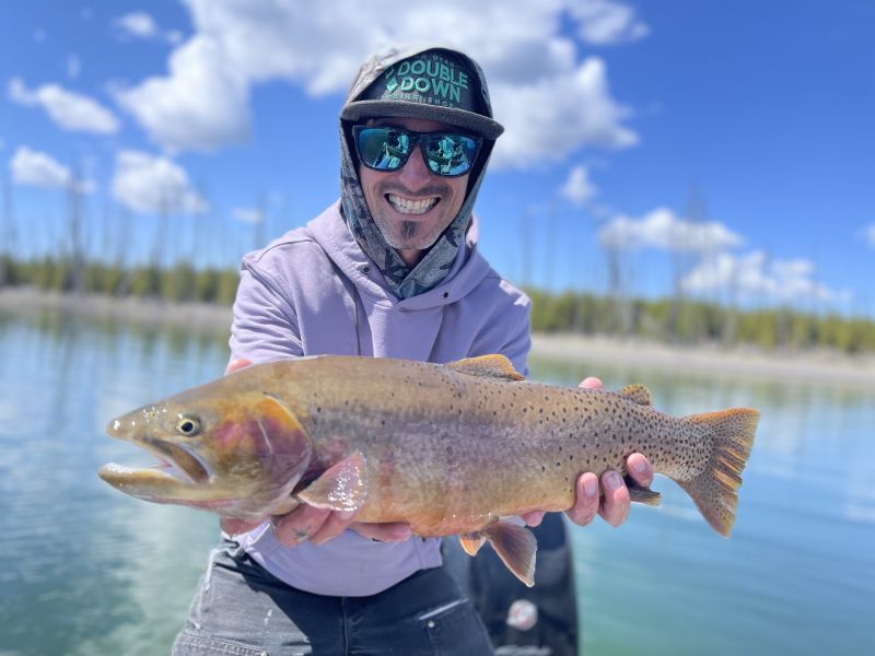 Angler holding a large trout on a boat, wearing sunglasses and a cap on a sunny lake.