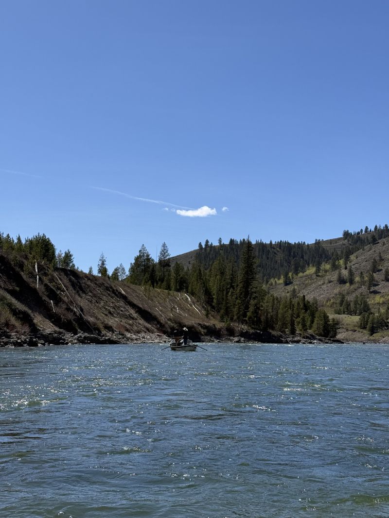 Rocky riverbank under a clear blue sky, with rippling water in the foreground.