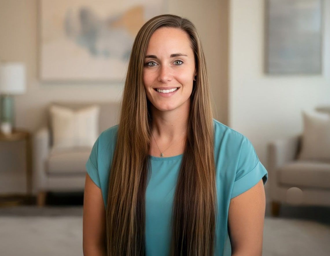 Woman with long brown hair smiling at the camera, wearing a teal top; indoor setting.