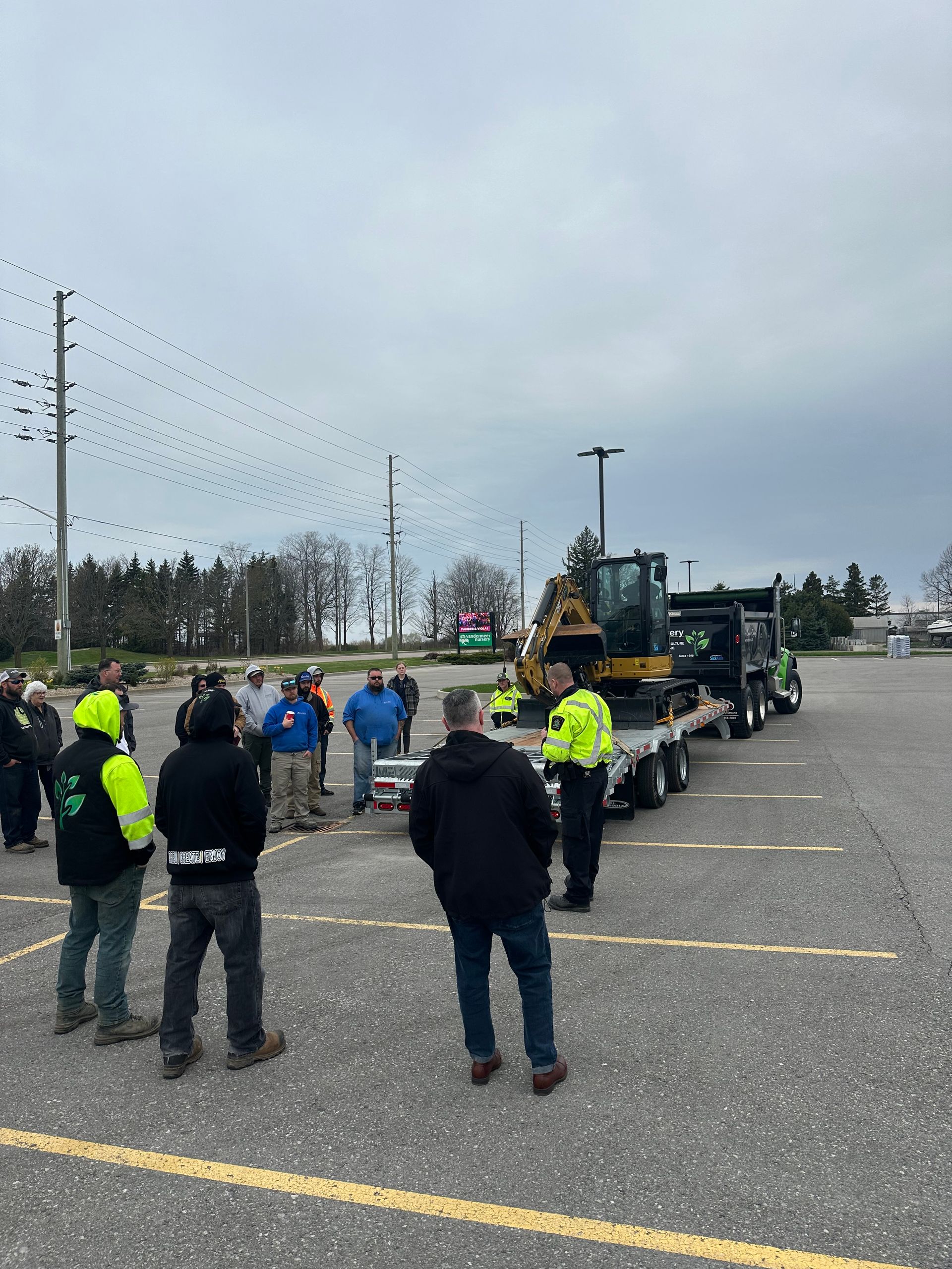 A group of men are standing in a parking lot watching a truck with a bulldozer on it.