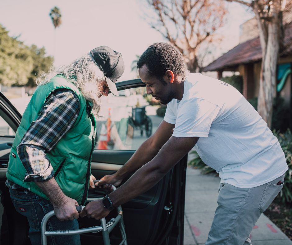 A man is helping an older man with a walker into a car.