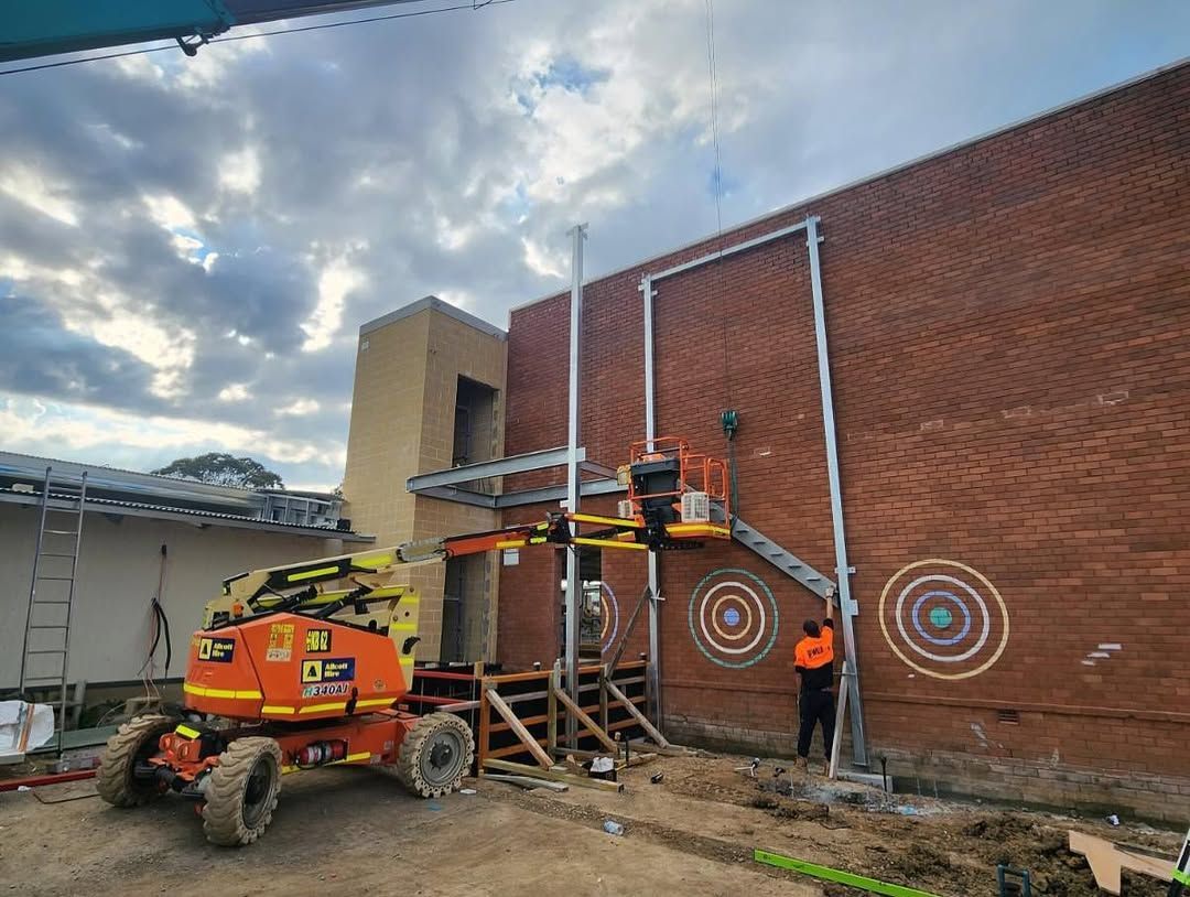 Construction Workers on a Brick Building Using a Lift to Install Metal Framework — Proweld Steel Fabrication In Unanderra, NSW