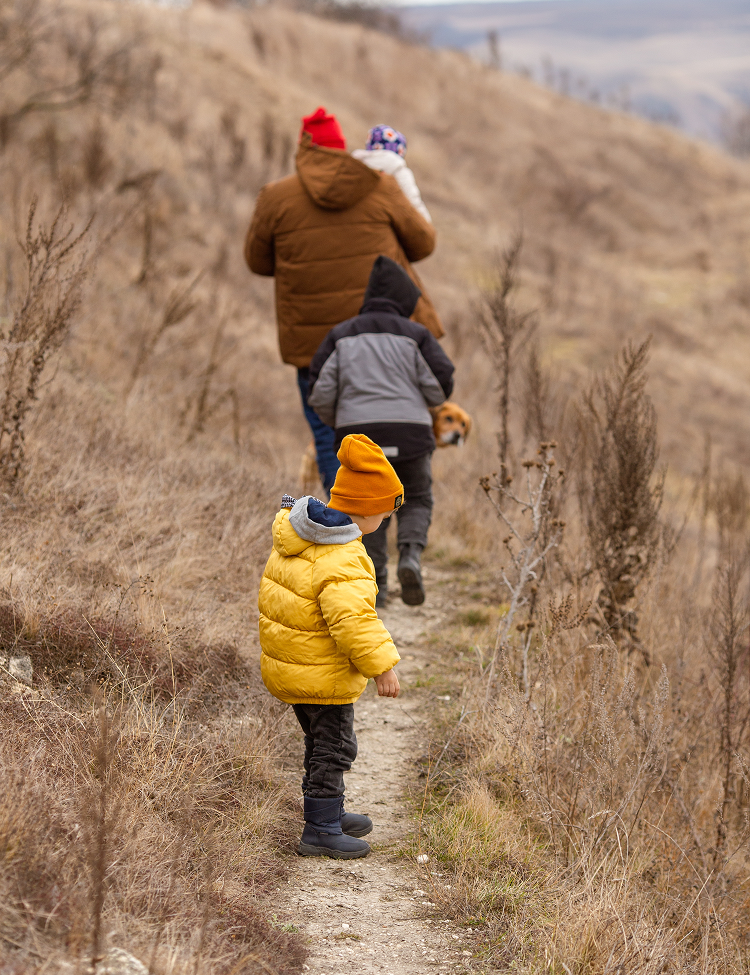 A family hiking on a trail. A child in a yellow jacket leads, followed by a group of people in winter clothes.