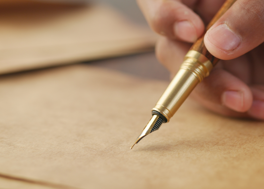 Hand holding a gold-tipped fountain pen writing on tan paper.