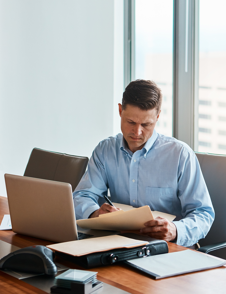 Man in a blue shirt at a desk writing on a file, with a laptop and phone, indoors.