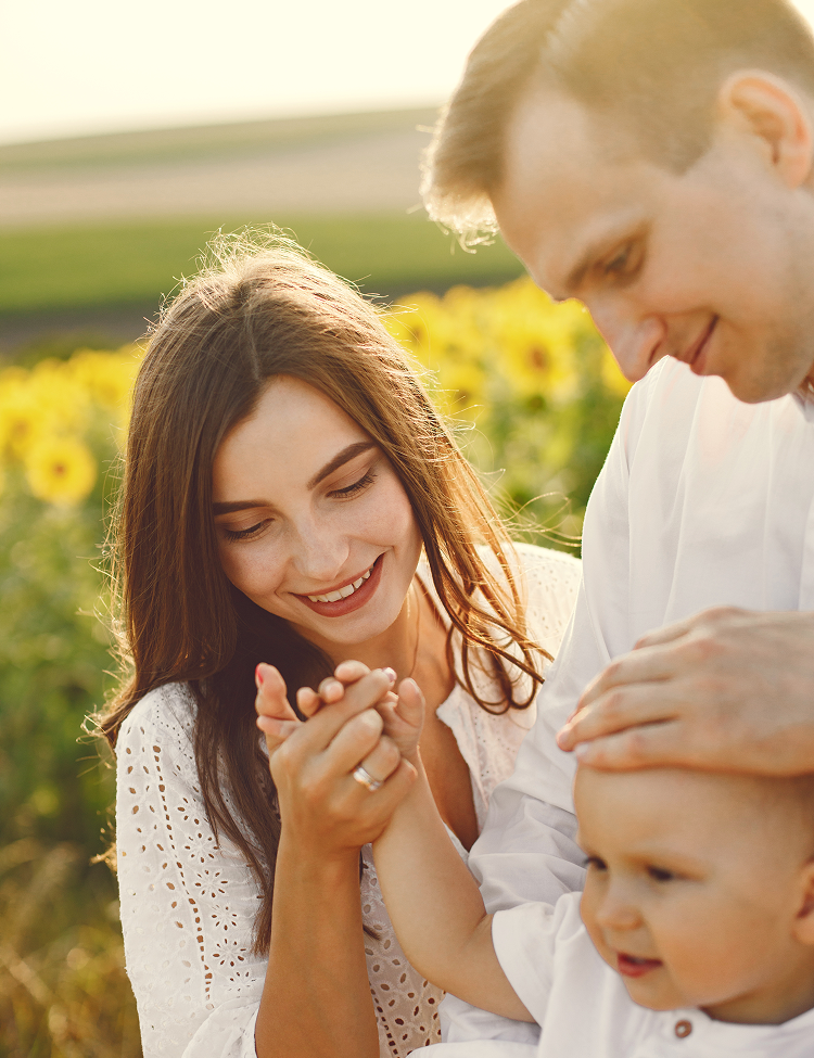 Family of three in a sunflower field: woman holding baby's hand, man touching baby's head, all smiling.