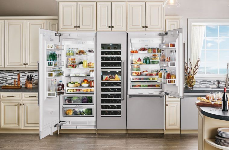 Stainless steel built-in refrigerator and wine cooler with open doors, filled with food, in a kitchen with beige cabinets.