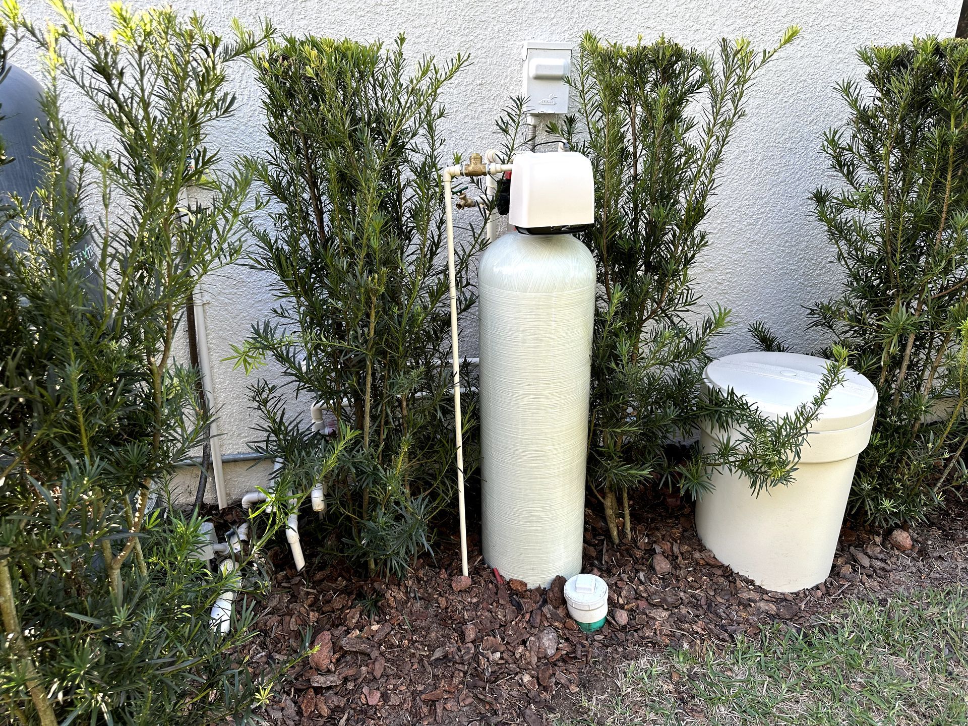 A water softener is sitting in the grass next to a bush.