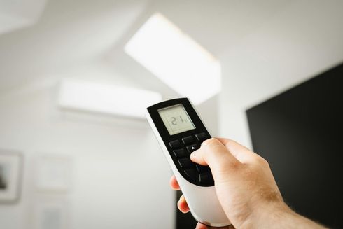 Hand holding AC remote, adjusting settings. Air conditioner visible in white room.