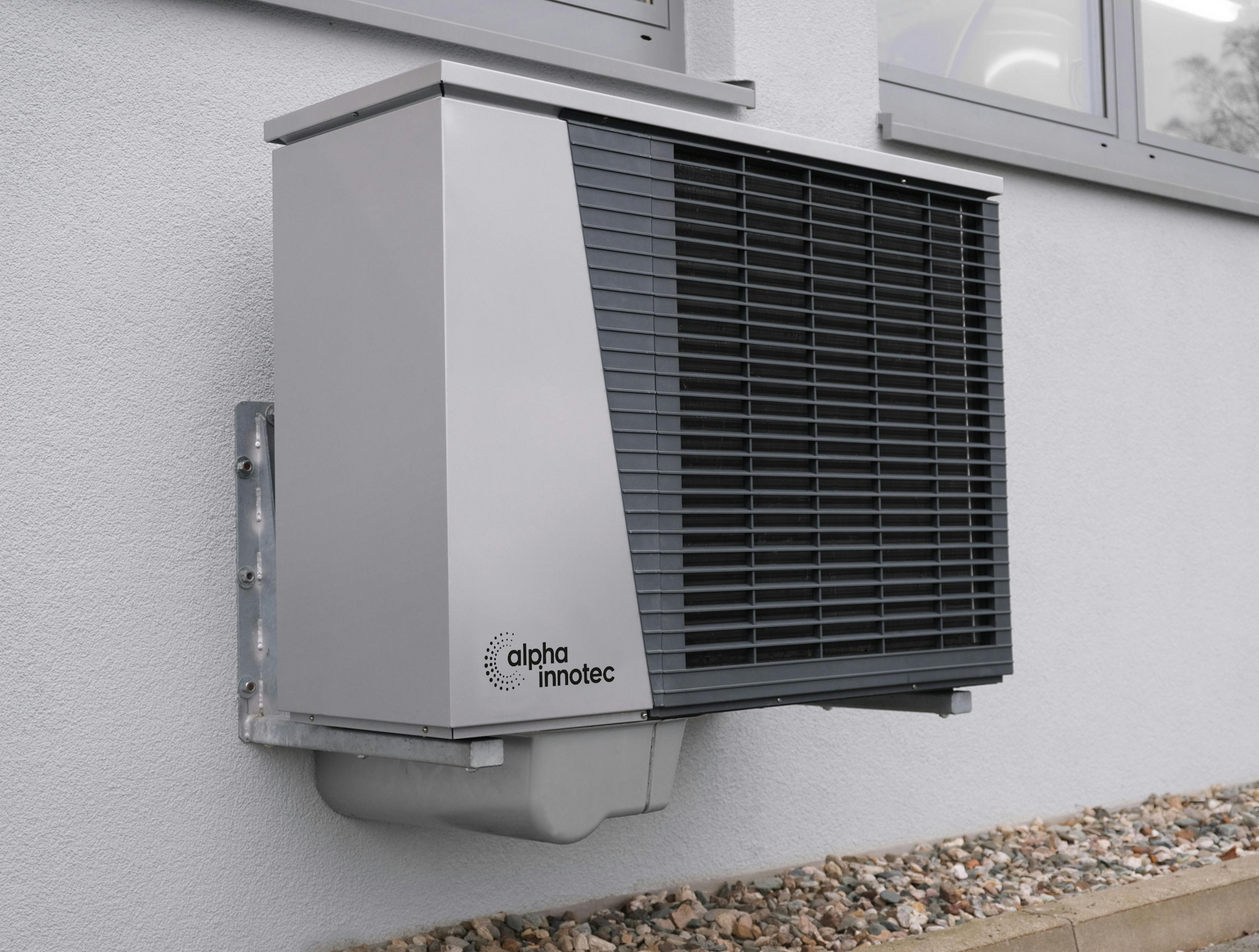 A wall-mounted, gray, rectangular air conditioning unit with black grille, mounted on a white building wall.