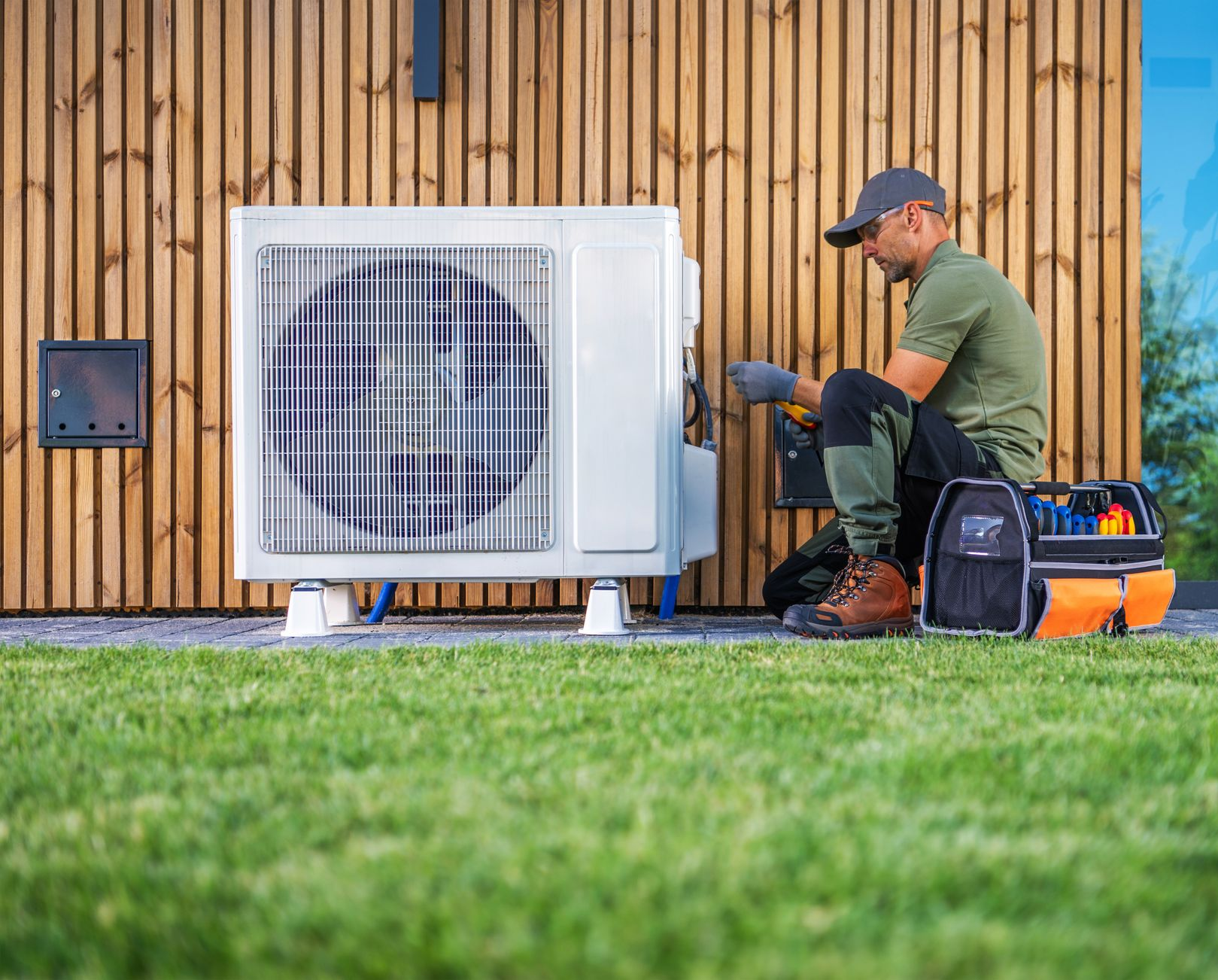 Man in work clothes repairs a heat pump unit outside a building with wooden siding, next to a toolbox.