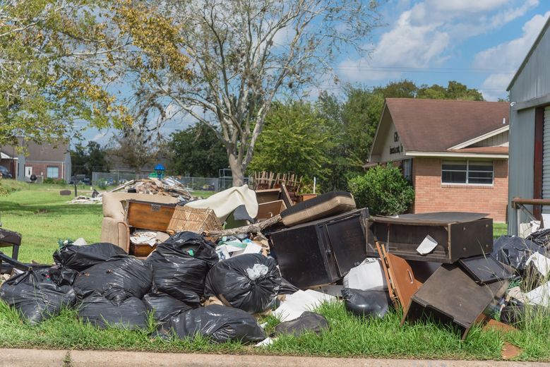 Pile of garbage bags, furniture, and debris on a curb near a house and trees.