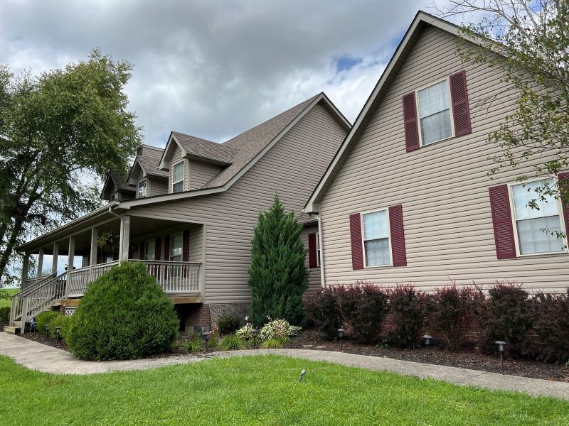 Tan two-story house with brown roof and red shutters. A covered porch extends to the left. Lush green grass and shrubbery in front.