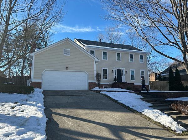 Beige two-story house with attached garage. Snow on the ground and driveway. Person walking by the house.