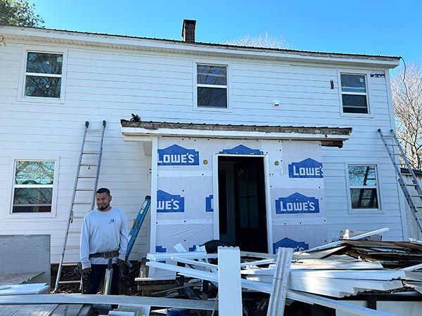 Man stands in front of a house under construction. The siding is white, windows are installed and the entrance is covered in Lowe's branded materials.