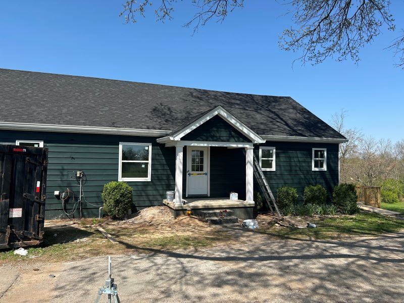 A dark green house with a newly painted white door and windows. A black dumpster sits on the left.