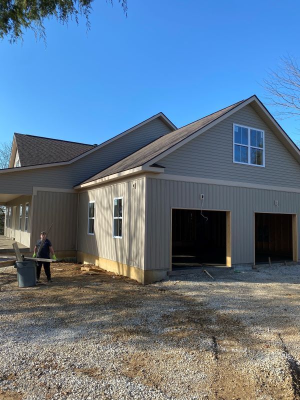 New house under construction with two garage bays, beige siding, and a clear blue sky. A person stands by a trash can.