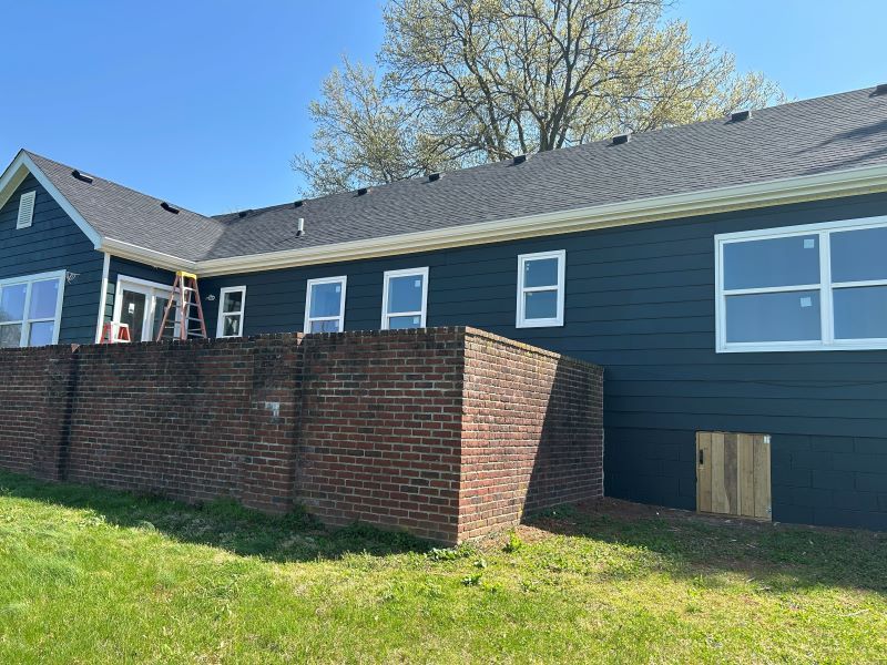 A brick wall in front of a blue house with white-framed windows under a blue sky.