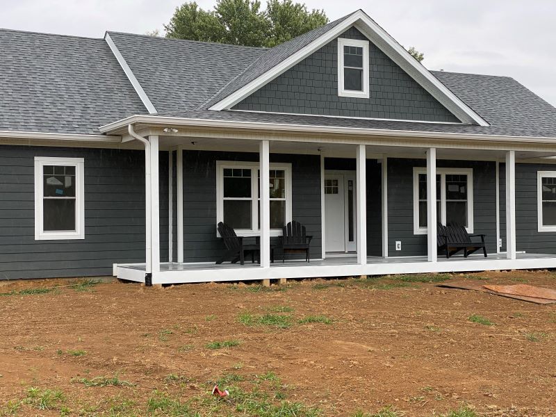 A newly built, gray house with white trim and a covered porch, set on a brown dirt lot.