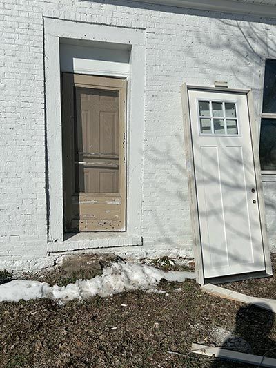 Two white doors, one standing, one removed. A building's white-painted brick exterior shows the empty doorframe.