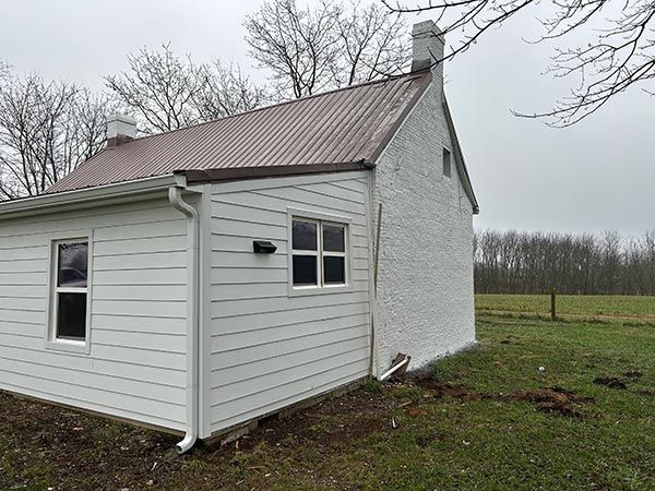 White building with brown metal roof and chimney.  It has white siding on one side and textured white stucco on the other, set in a grassy field.