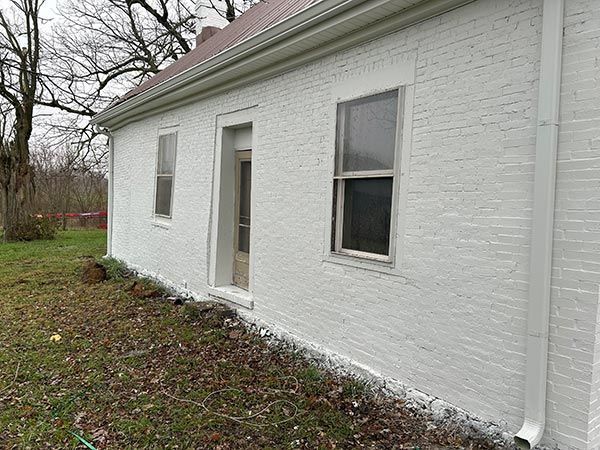 A white brick house with a brown metal roof and a white downspout. The side of the house has two windows and a doorway.