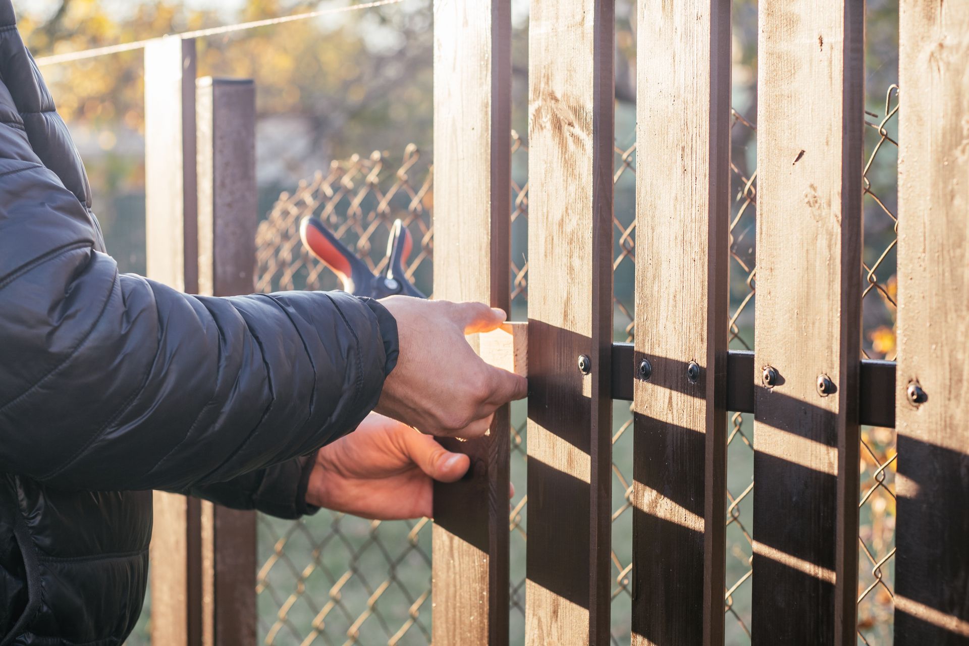 Person repairing a wooden fence with a pair of pliers outside on a sunny day.
