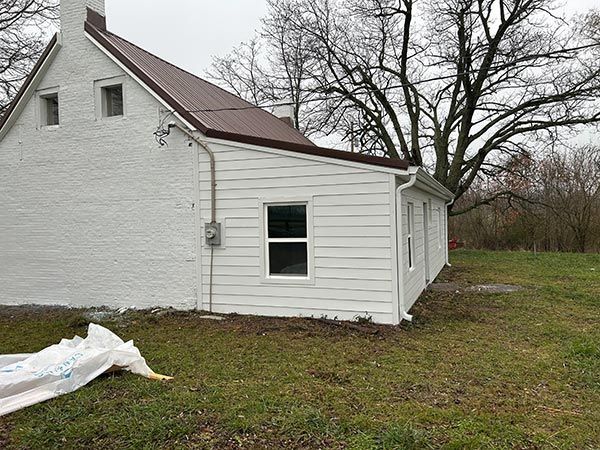 White house with brown roof and siding on a grassy lawn. A small window and a power meter are visible. Overcast sky.