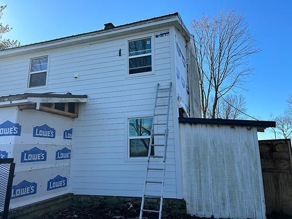 Two-story house under renovation, with new white siding. A ladder rests against the side, and Lowe's house wrap is visible.