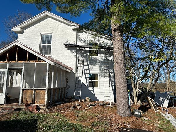 White house with a screened porch undergoing renovations. Ladders lean against the building, debris scattered on the ground.