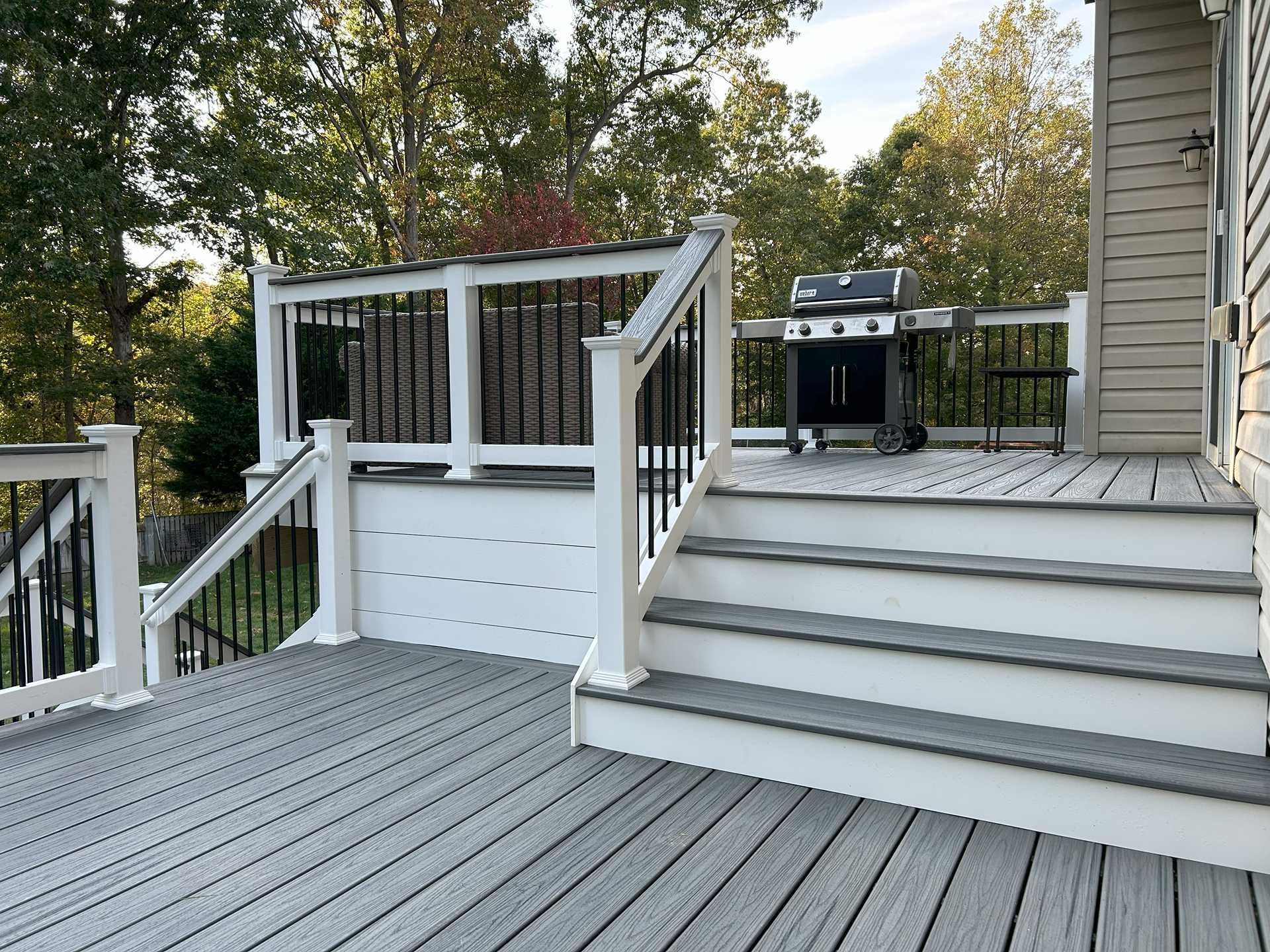 Gray deck with stairs and grill, white and black railings, surrounded by trees.