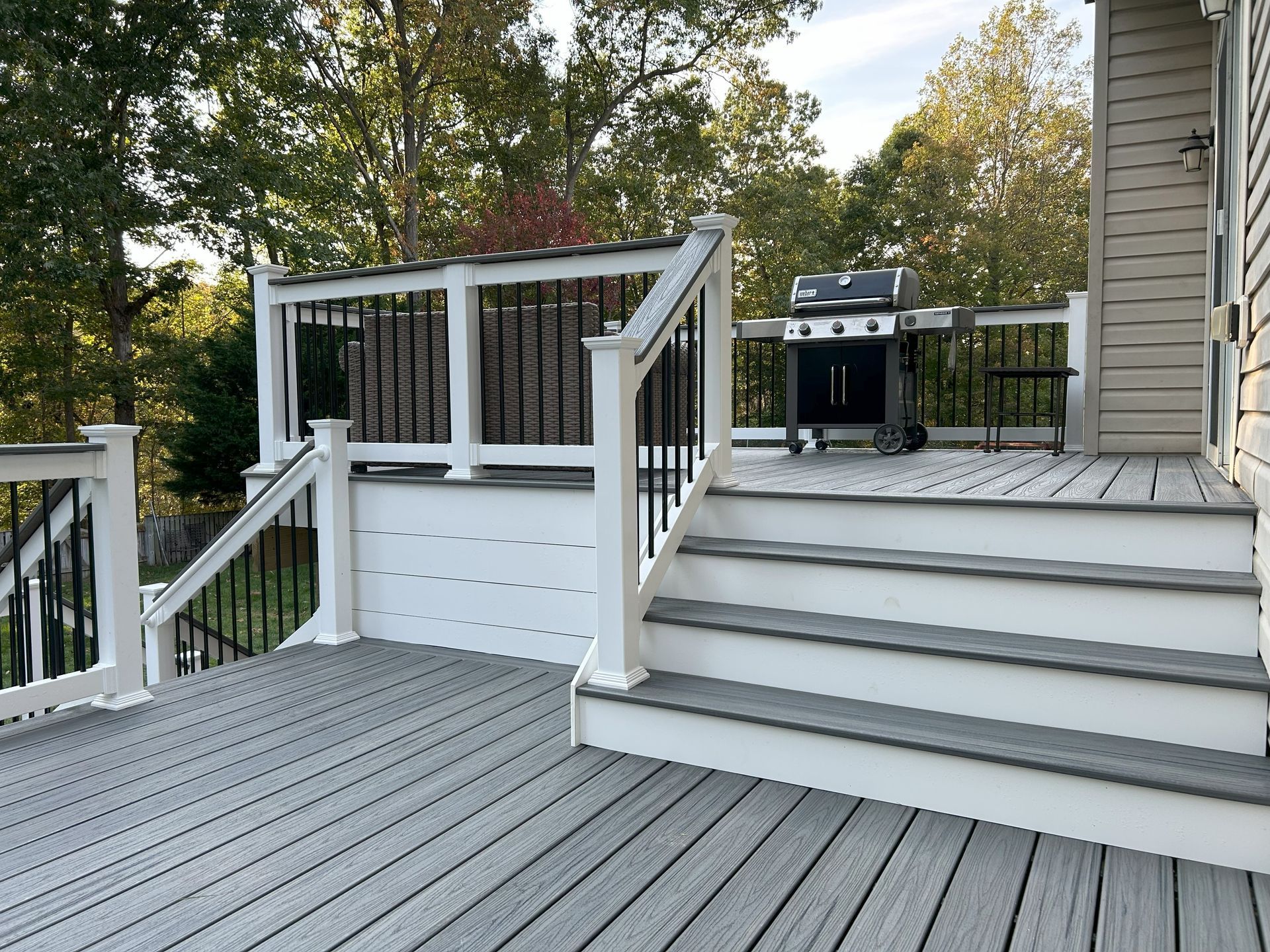 Gray deck with white railing and stairs; black grill sits on the deck, surrounded by trees.
