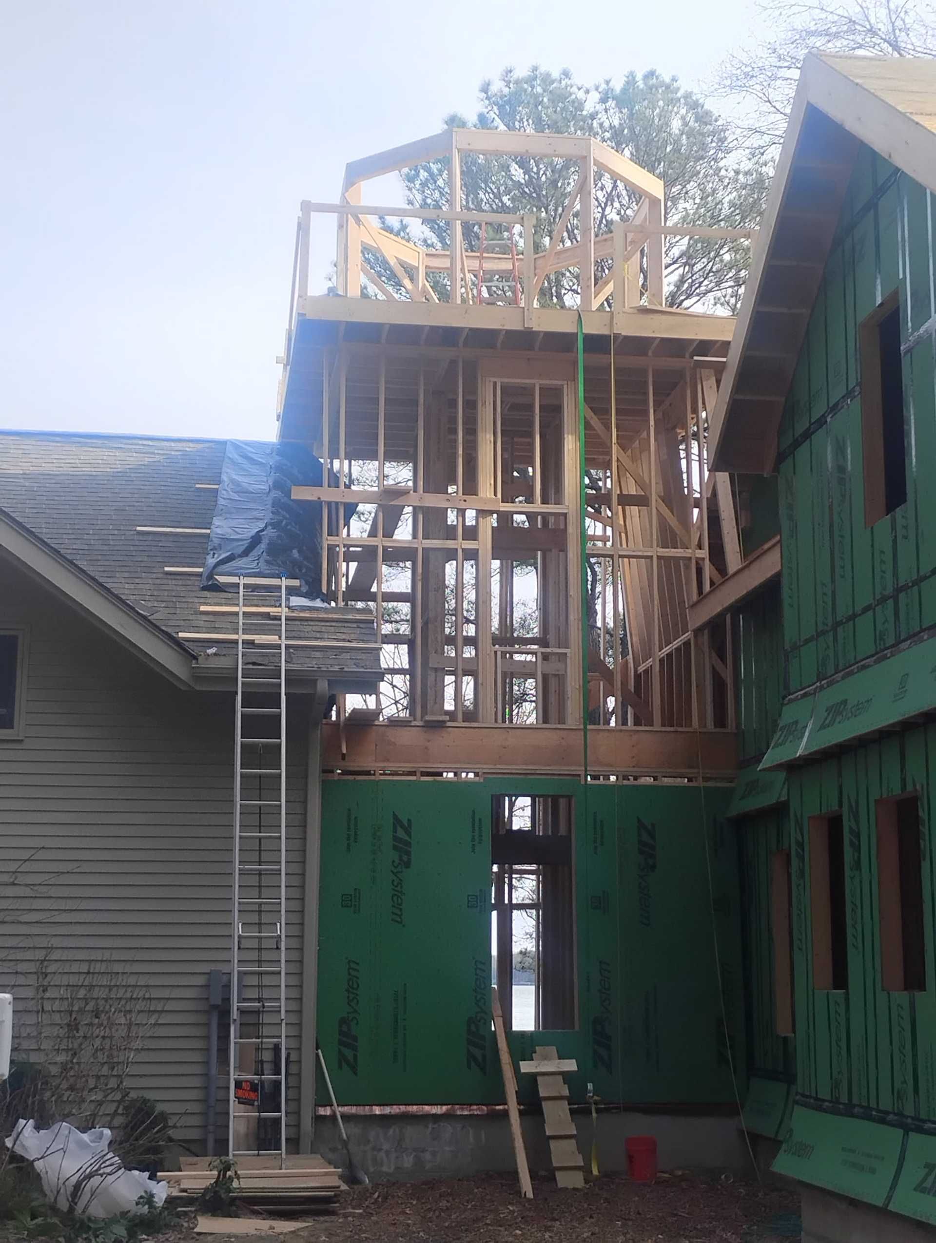 Construction site: framing of a two-story addition to a house. Green sheathing and wooden beams are visible.