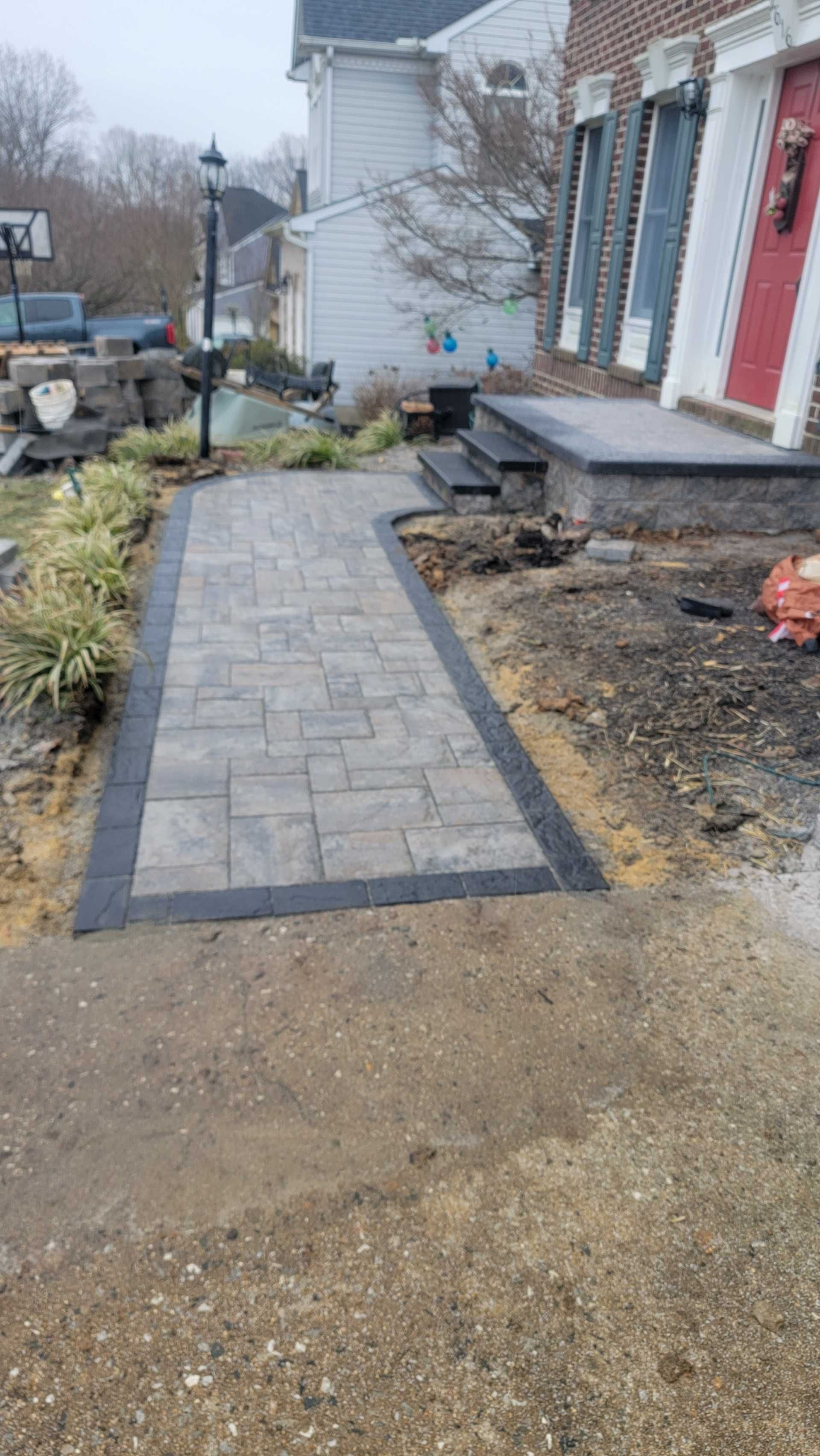 Brick walkway leading to a house with a red door. Black border and landscaping on the sides.