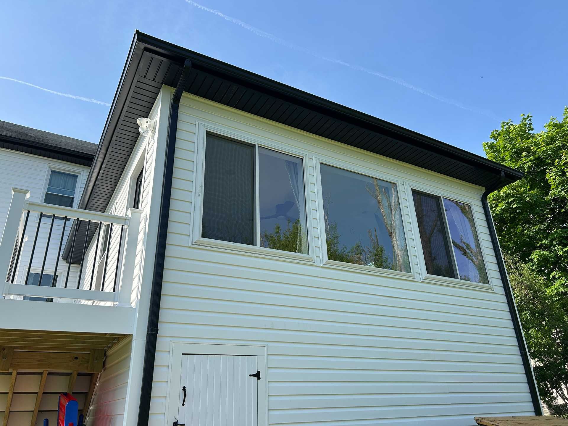 White siding sunroom with black roof and windows, attached to a house with a deck.