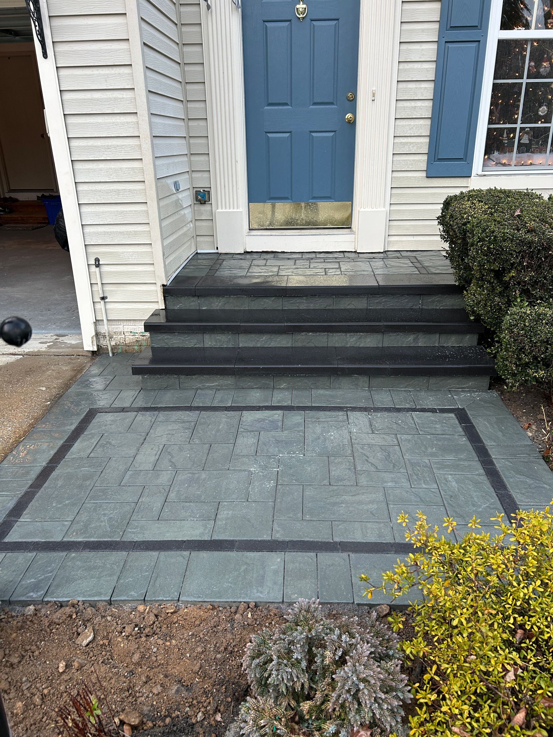 Gray brick walkway and steps leading to a blue door. Black border and shrubs.