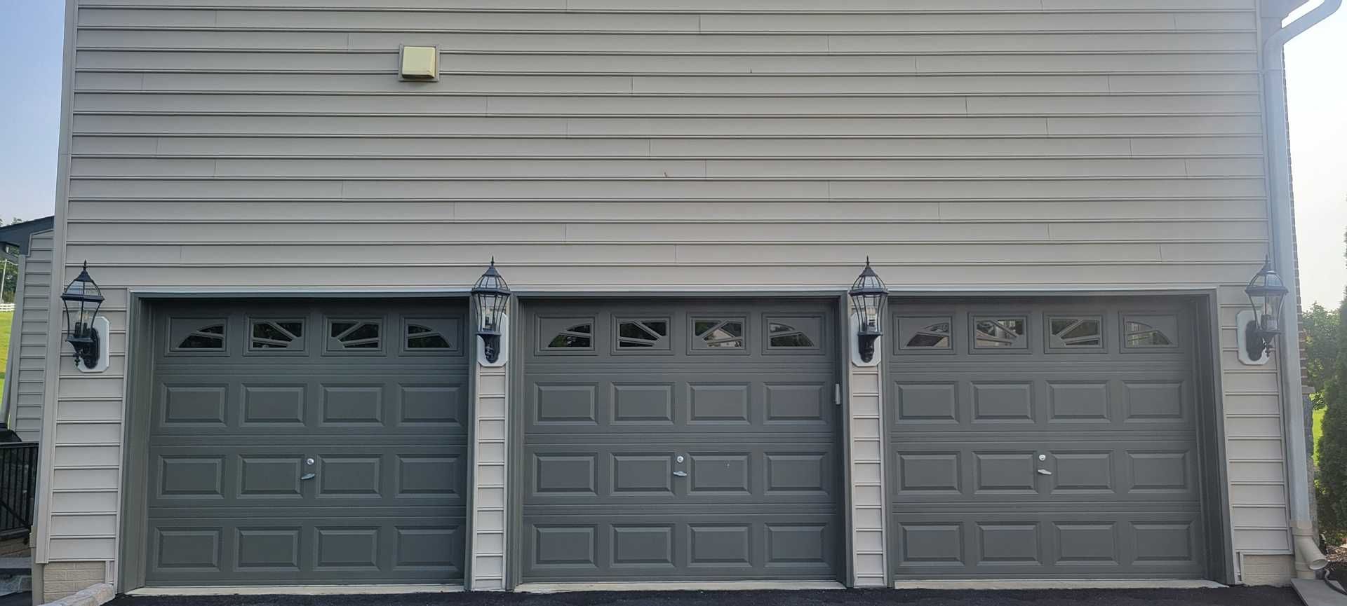Three gray garage doors with matching wall siding, black sconces, and a blue sky.