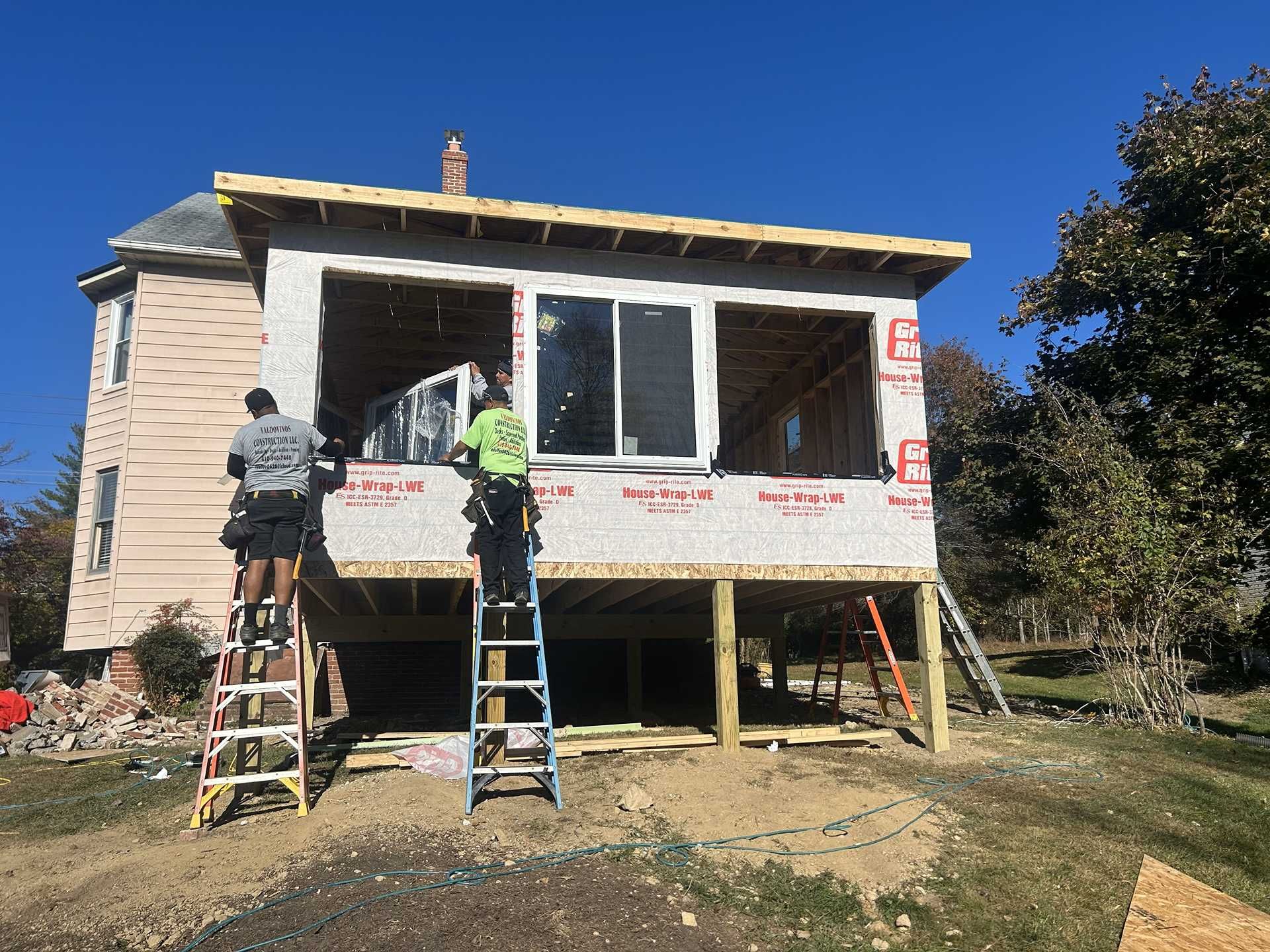 Construction workers building an addition to a house. Two workers on ladders, framing windows, bright sunny day.