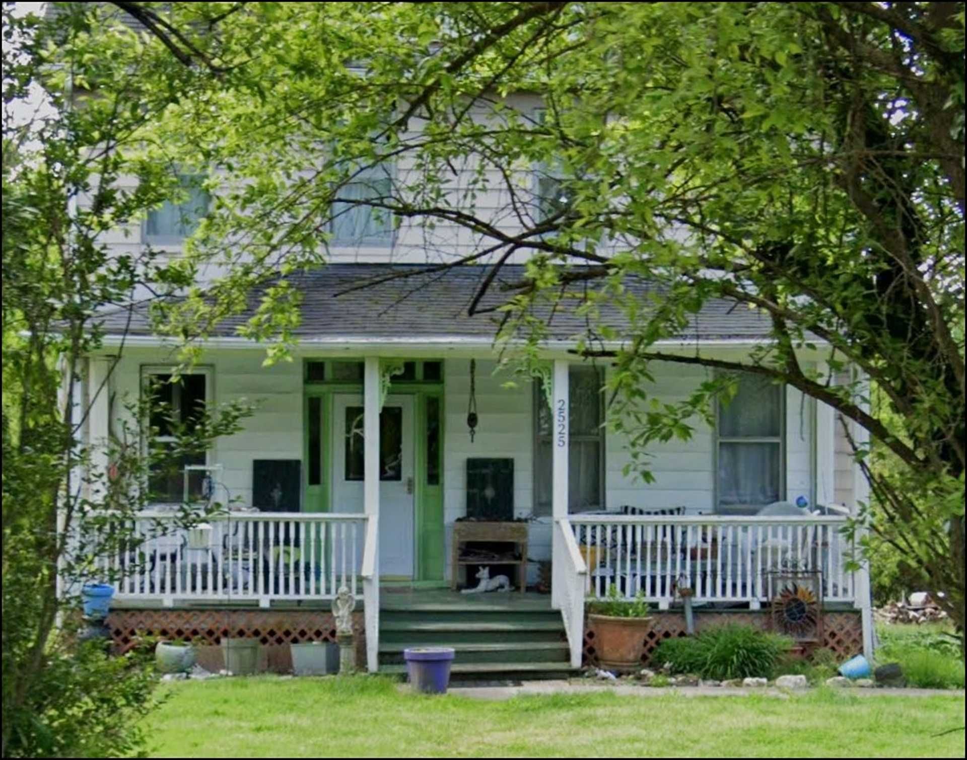 White two-story house with green accents and a porch, framed by leafy green trees.