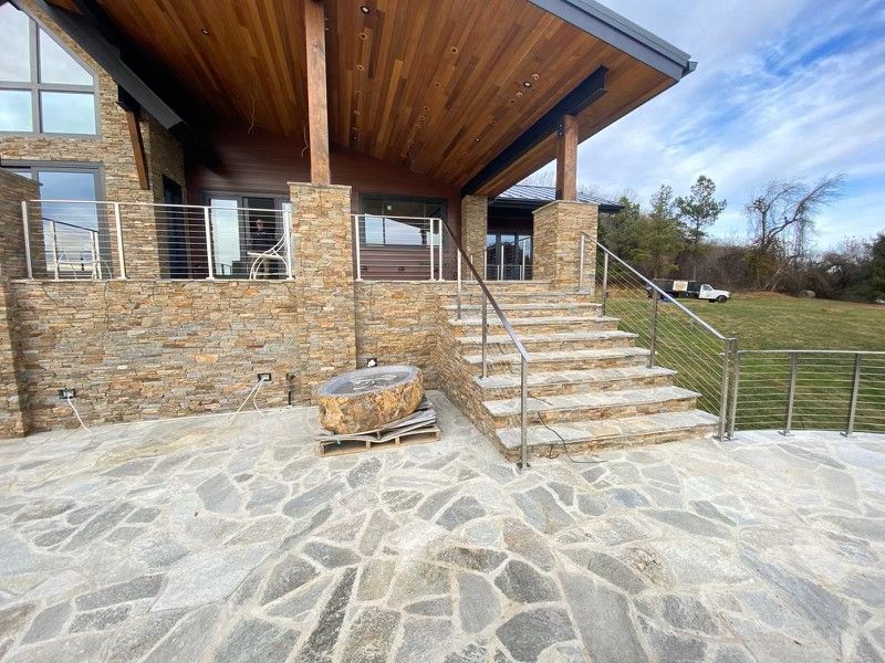 Stone-clad house exterior with a patio, steps, and handrails. Wooden ceiling and lawn in the background.
