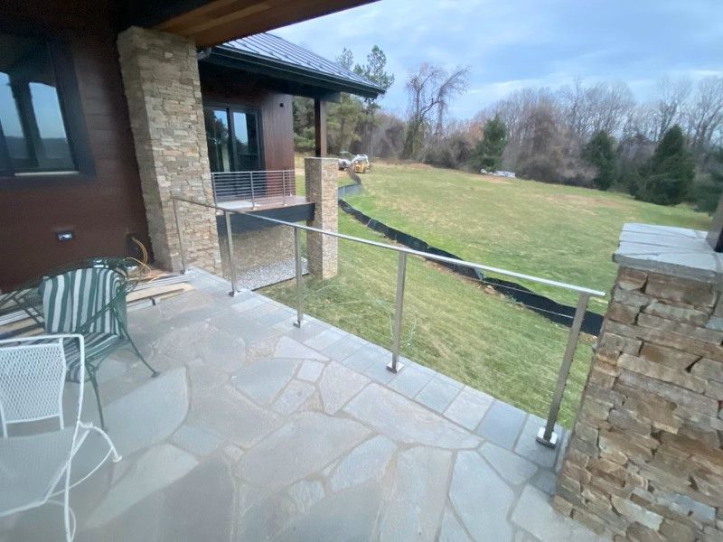 Patio with stone floor, stainless steel railing, and grassy yard beyond. A house with stone columns is on the left.