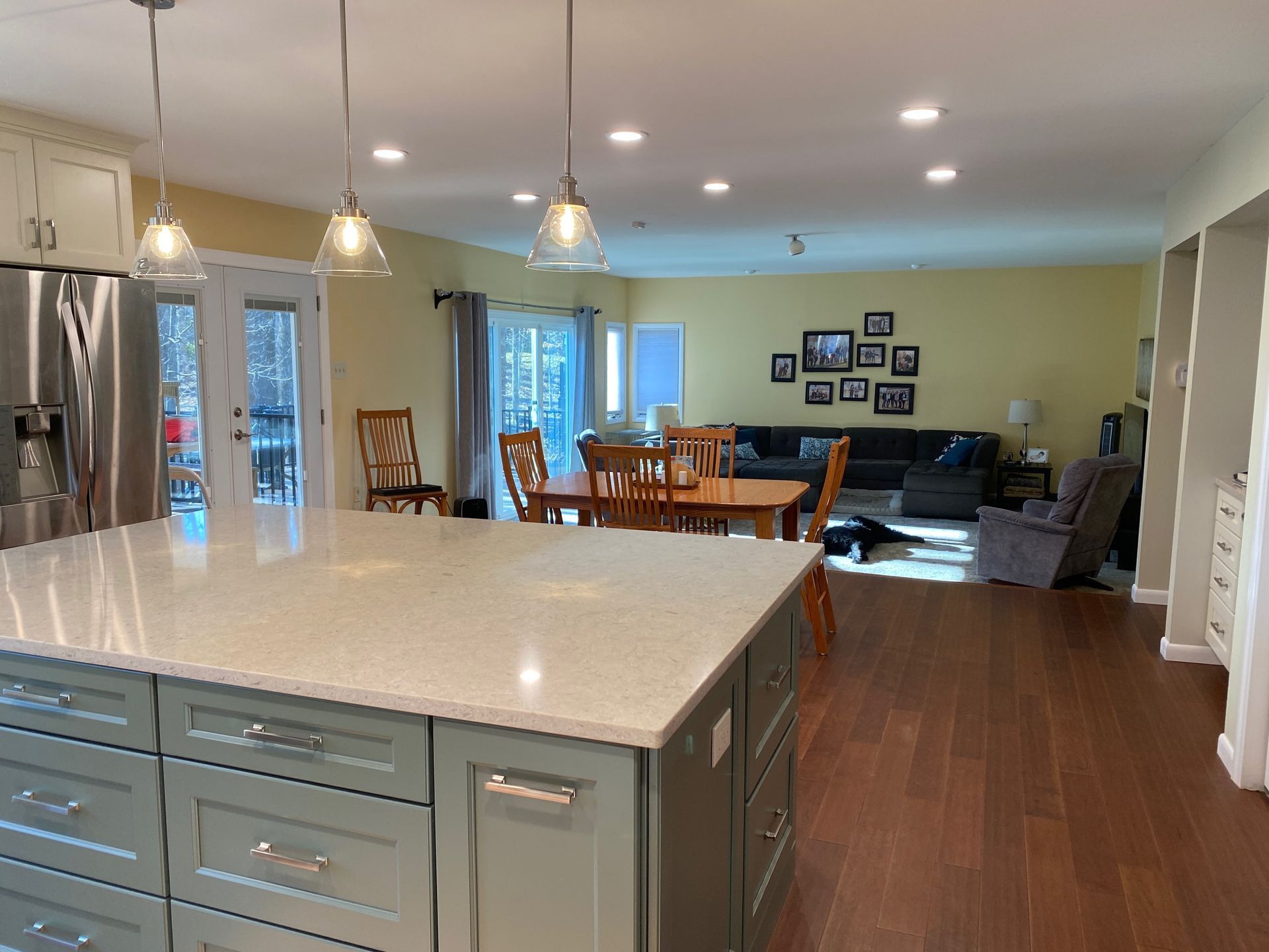 Spacious kitchen with island, dining area, and living room. Light green cabinets, wood floors, yellow walls.