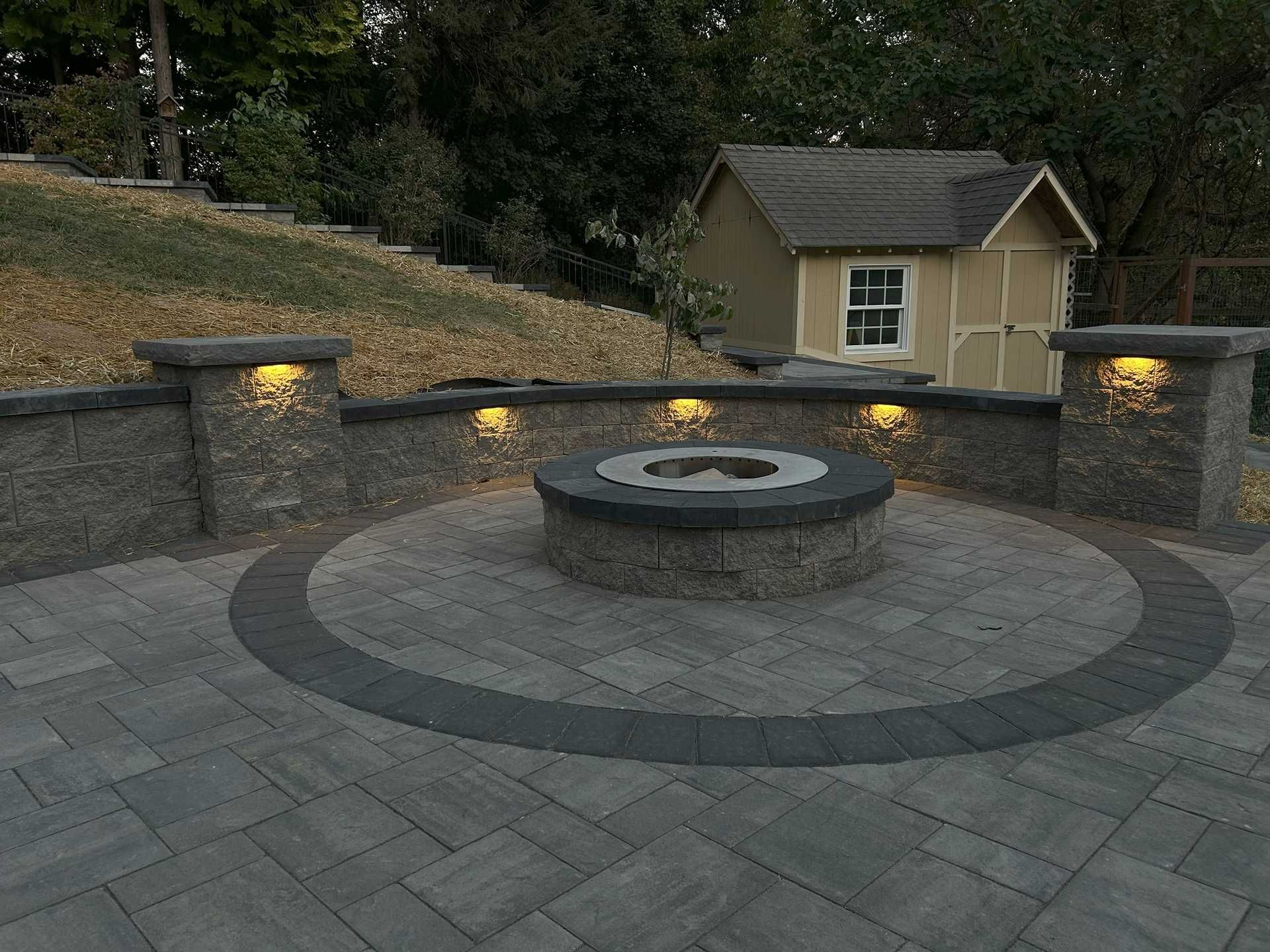 Stone patio with fire pit, illuminated wall, small shed in the background.