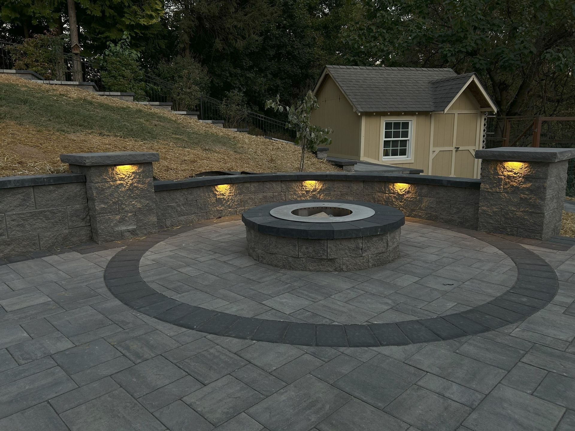 Outdoor patio with fire pit, stone walls, and ambient lighting in a yard with a shed in the background.