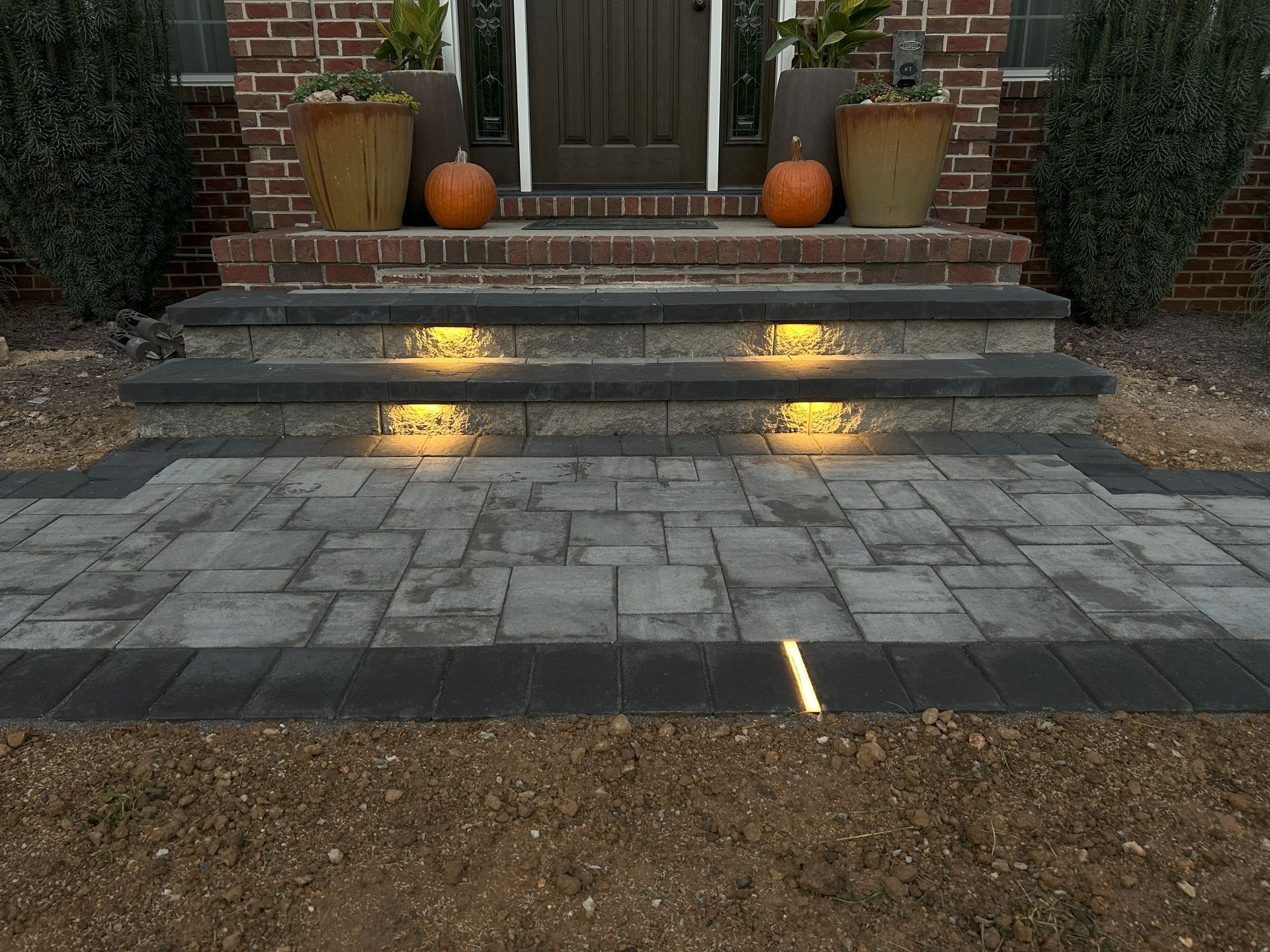 Brick pathway and steps leading to a front door, with pumpkins and potted plants for decoration. Step lights are illuminated.