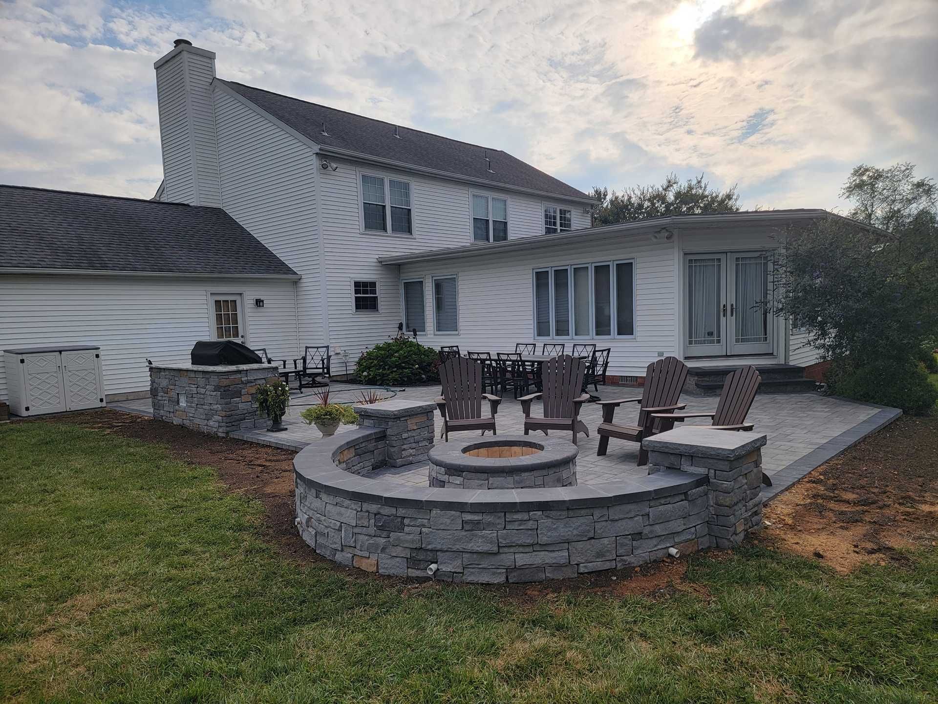Backyard patio with fire pit, seating, and a two-story white house with a partially visible overcast sky.
